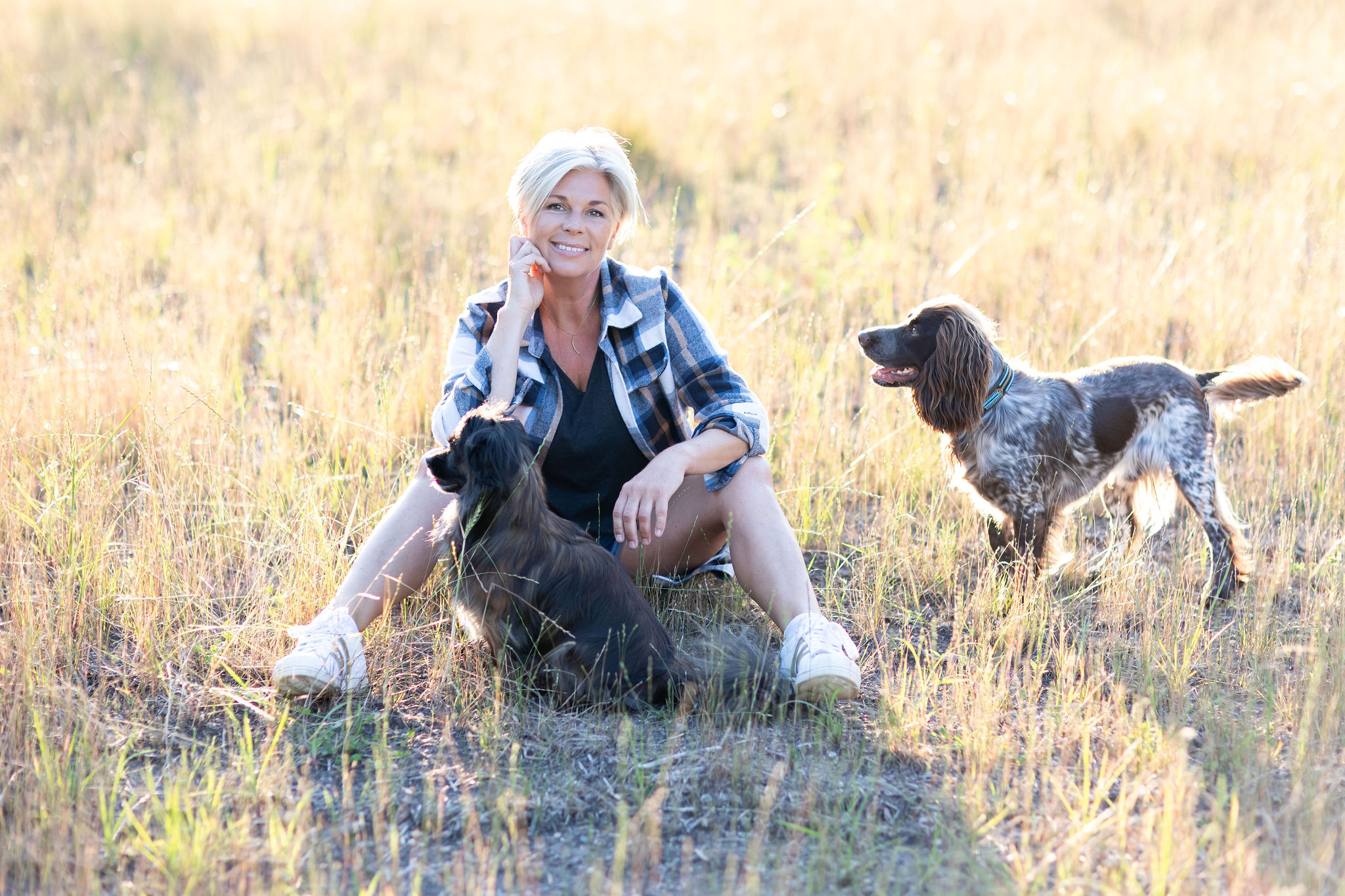 Justine Schuurmans sitting in a field with two dogs, one lying beside her and the other standing nearby, in a setting bathed in soft sunlight.